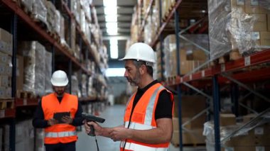 Lojistik müdürü, büyük bir dağıtım merkezi koridorundaki stokları kontrol etmek ve yönetmek için barkod tarayıcısının ve tabletin nasıl kullanılacağı konusunda yeni bir çalışanı denetliyor ve eğitiyor.