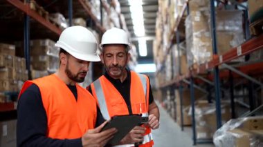 İki depo çalışanı envanteri yönetmek için işbirliği yapıyor, biri kutularda barkod tarayıcısı kullanıyor ve diğeri de uzun raflardaki stokları kontrol ederken dijital tablete veri giriyor.