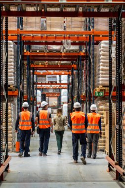 Lojistik müdürü, modern bir dağıtım merkezinde güvenlik yelekleri ve sert şapkalar giyen bir depo çalışanı ekibiyle hisse senedi seviyelerini ve operasyonlarını inceliyor.