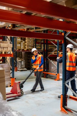 Depo çalışanlarından oluşan çeşitli bir ekip, bir dağıtım merkezindeki depolama raflarının arasında bir el paleti kamyonuyla mal taşıyarak ve envanterle uğraşırken iletişim kuruyor.