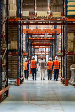 Çeşitli işçiler ve güvenlik yelekleri ve kasklı bir yönetici dolu modern lojistik deposundaki karton kutular arasında uzun bir koridorda yürüyorlar.