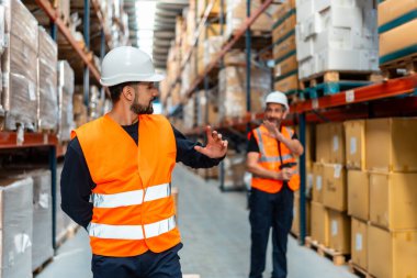 Sert şapkalı ve güvenlik yelekli iki erkek depo çalışanı yoğun bir dağıtım merkezi koridorunda envanteri tartışıyor üst raflarda istiflenmiş kutular, takım çalışmalarını ve lojistiği gösteriyor.