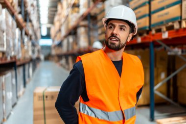 Sert şapka ve turuncu güvenlik yeleği giymiş bir depo işçisi koridorda duruyor, envanter ve kutuları inceliyor, modern bir dağıtım tesisinde stokları kontrol ediyor.