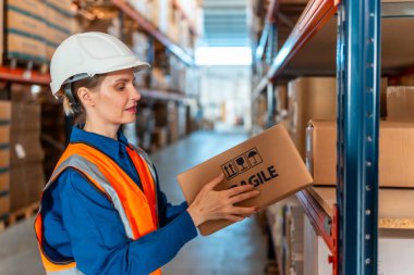 Güvenlik kaskı ve yelek giyen bir kadın depo çalışanı, modern bir lojistik deposunda güvenli depolama ve envanter yönetimi için hassas etiketli bir karton kutuyu inceliyor.