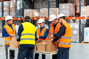 Muhtelif depo ekipleri kasketli ve çelik yelekli. Uzun bir masa üzerinde karton paketleme ve sıralama. Yoğun bir endüstriyel dağıtım merkezi içinde, verimli tatbikat operasyonlarına odaklanmış.