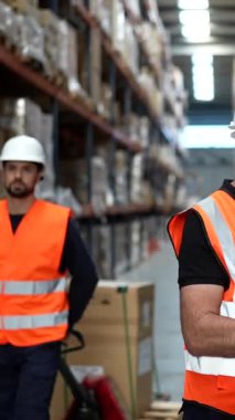 Envanter yönetimi için tarayıcı kullanan bir erkek depo çalışanı. Arka planda, büyük bir dağıtım merkezinde karton kutularla dolu rafların arasında duran bir meslektaşı var.