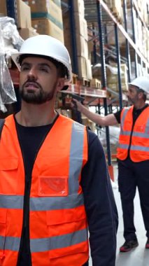 Depo yöneticisi ve kadın yöneticiye envanter kontrolü sırasında yardım eden bir çalışan, büyük bir dağıtım merkezi koridorundaki raflardaki stokları yönetmek için barkod tarayıcıları kullanan işçiler.