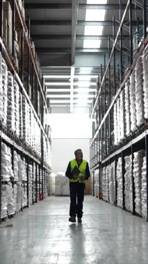 Yüksek görüş mesafesi olan bir yeleği olan kendinden emin bir erkek depo çalışanı elinde bir pano ve tarayıcı ile büyük raflardaki envanterleri kontrol ederken büyük bir depo koridorunda yürüyor.
