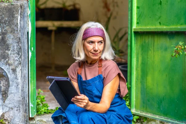 Yeşil bir binanın dışında dijital tablet üzerinde çalışan önlüklü ve bantlı kıdemli kadın sanatçı, yaratıcı projesini çizerken ve planlarken düşünceli ve odaklanmış.