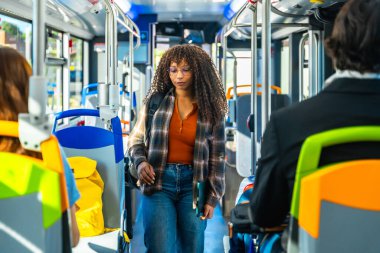 Uzun kıvırcık saçlı ve gözlüklü genç bir kadın otobüs koridorunda yürüyor, dizüstü bilgisayar ve sırt çantası taşıyor, içi parlak koltuk renkleriyle dolu.