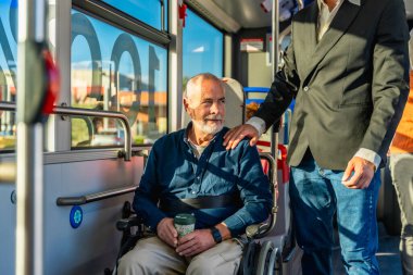 Toplu taşıma araçlarını kullanan yaşlı bir adam, bir otobüste tekerlekli sandalyesinde otururken ilgili bir asistandan yardım ve duygusal destek alıyor.