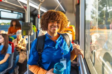 Mavi önlüklü ve sırt çantalı genç hemşire, bir belediye otobüsünde dururken kameraya gülümsüyor, işe gidiş gelişi sırasında önemli bir sağlık çalışanı olarak tekrar kullanılabilir bir bardak tutuyor.