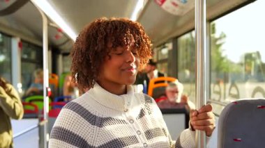 Otobüsün içinde durup kameraya gülümseyen kıvırcık saçlı neşeli Afro-Amerikalı kadın. Toplu taşıma yolcusu güneşli bir günde şehirde günlük seyahatinin keyfini çıkarıyor.