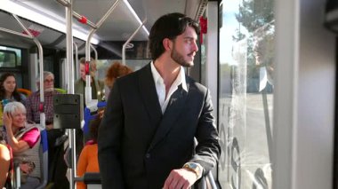 Kamu otobüsünün içinde duran koyu renk takım elbiseli yakışıklı bir profesyonel. Düşünceli bir şekilde pencereden dışarı bakıyor. Şehir içi seyahati sırasında diğer yolcularla işe gidip geliyor.