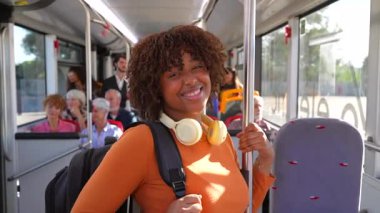 Kıvırcık saçlı ve sırt çantalı güzel bir Afro-Amerikan kadının portresi. Şehir otobüsünde giderken kameraya gülümsüyor. Günlük yolculuğu boyunca tırabzanlara tutunuyor.