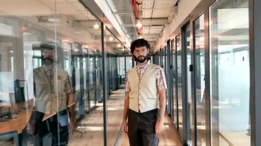 Ofis koridorunda yürüyen, kollarını kavuşturup kameraya gülümseyen, kariyerinin başarısını ve memnuniyetini yansıtan sakallı ve gözlüklü, kendine güvenen genç bir profesyonel.