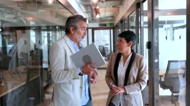 Olgun iş ortakları birlikte yürüyorlar ve cam duvarlı koridor boyunca profesyonel bir sohbete giriyorlar. Çağdaş bir çalışma alanında işbirliği ve takım çalışmasını gösteriyorlar.