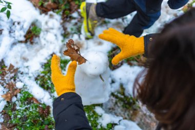 Sarı yün eldiven giyen çocuk, karlı bir ormanda kahverengi yapraklı küçük bir kardan adamı süslüyor. Üst manzarayı kapatıyorlar ve neşeli kış yaratıcılığını ve açık hava eğlencesini gösteriyor.