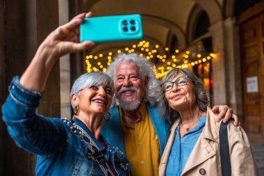 Üç mutlu kıdemli arkadaş şehir gezisinde eğleniyor, gülümsüyor ve kucaklaşıyorlar, akıllı bir telefonla neşeli bir selfie çekiyorlar, bir anlık dostluk ve seyahat yakalıyorlar.