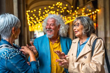 Dekoratif ışıklar altında canlı sohbetin keyfini çıkaran üç kıdemli arkadaş, bir şehir ortamında sosyalleşirken gülüşmeler ve pozitif enerjiyi paylaşıyorlar.