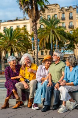 Güneşli bir günde palmiye ağaçlarıyla çevrili bir şehir meydanında, arkadaşlığı, emekliliği ve seyahati temsil eden bir bankta otururken gülümseyen ve etkileşen bir grup arkadaş.