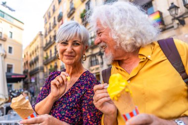 Mutlu, olgun bir çift, dondurma yerken birlikte gülümsüyor, bir şehir tatili sırasında boş vakit ve aşk yaşıyor, arka planda kapsayıcılığı temsil eden bir gurur bayrağı var.