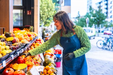 Gülümseyen manav kadın önlük ve eldiven takıyor. Küçük işletmesinin dışındaki marketlerde taze meyve ve sebze düzenliyor. Kaliteyi ve sunumu garantiliyor.