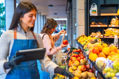 Gülümseyen ve elinde dijital tablet tutan bir manav. Bu sırada mutlu bir kadın tüketici açık hava pazarından taze domates, meyve ve sebze seçiyor. Sağlıklı beslenmeyi ve yerel işletmeleri teşvik ediyor.