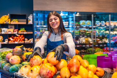 Üretimde taze nar, mango ve üzüm düzenleyen önlüklü ve eldivenli gülümseyen genç süpermarket çalışanı, kaliteyi, özeni ve sağlıklı perakende hizmeti vurguluyor