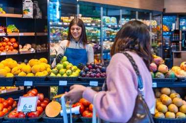 Dost canlısı bir süpermarket çalışanı, önlük ve eldiven giyer. Üretim bölümünde çalışan bir kadına taze yeşil elma sunarak yardımcı olur. İyi hizmet ve sağlıklı yemek seçeneklerini ön plana çıkarır.
