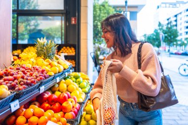 Gülümseyen bir kadın modern bir süpermarketteki vitrinden taze organik meyve seçiyor ve eko dostu iplik alışveriş çantasına ürünler yerleştiriyor.