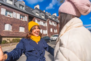 Kış kıyafetleri giymiş iki neşeli kadın bir şehir caddesinde kollarını açıp birbirlerini selamlıyor ve mutluluğu ve bağlantıyı ifade ediyorlar.