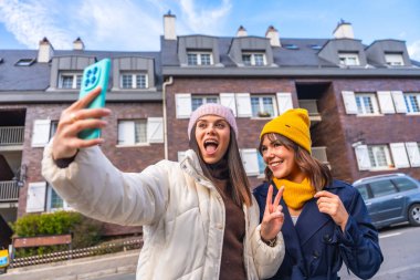 İki neşeli genç kadın, bir şehir caddesinde güneşli bir kış selfie 'si çekiyorlar. Bereler ve ceketlerle sarmalanmış, gülümsüyor ve birlikte eğlenceli bir seyahat anı yakalıyorlar.