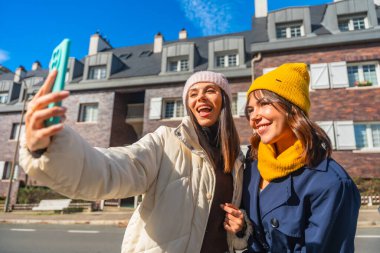 İki neşeli genç bayan arkadaş şehirde güneşli bir kış gününün tadını çıkarıyorlar, gülümsüyorlar ve akıllı bir telefonla eğlenceli bir selfie çekiyorlar, neşeli bir arkadaşlığı paylaşıyorlar.