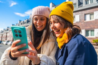 Kışlık şapkalar ve kalın giysiler giymiş iki genç kadın, aydınlık bir günde birlikte cep telefonuna bakarken gülüyorlar, neşeli bir anı paylaşıyorlar.
