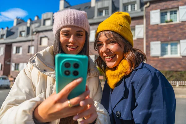 Kışlık ceketli ve bereli iki arkadaş gülümsüyor ve güneşli bir şehirde selfie çekiyor, samimi bağlantıların keyfini çıkarıyorlar, sosyal medya eğlencesi ve açık hava arkadaşlığı.