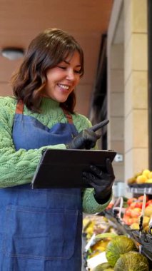 Meyve ve sebze dükkanında çalışan gülümseyen genç bir kadın stok ve siparişleri yönetmek için dijital tablet kullanıyor. Ürünlerin yanında dururken hijyen ve güvenlik için önlük ve eldiven giyiyorlar.