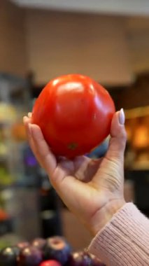 Genç bayan müşteri, süpermarketin taze üretim bölümünden büyük, olgun, kırmızı bir domates seçiyor ve onaylanması ya da tartılması için gülümseyen bir kadın manava gösteriyor.