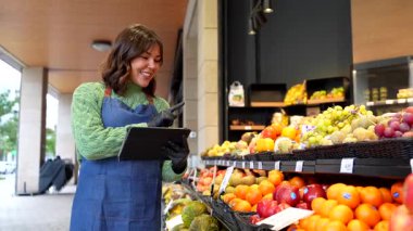 Bir meyve ve sebze dükkanının önünde dikilirken dijital tablet kullanan önlüklü genç bir kadın, küçük işletmelerde modern teknoloji sergiliyor.
