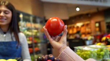 Koruyucu önlüklü ve eldivenli arkadaş canlısı manav, modern bir marketin veya süpermarketin taze üretim bölümünden olgun, kırmızı domates seçmesine yardım ediyor.