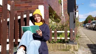 Sarı bereli ve mavi trençkotlu güzel genç bir kadın güneşli bir sonbahar gününün tadını çıkarıyor, bir şehir caddesinde ahşap bir çitin kenarında oturuyor ve kitap okurken gülümsüyor.