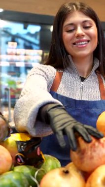 Önlüklü ve eldivenli gülümseyen genç bir kadın meyve vitrininde taze narı dikkatlice düzenliyor, mutlu ve olumlu bir tavırla modern bir markette gayretle çalışıyor.