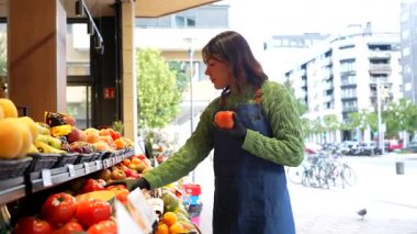 Genç bayan satıcı, yerel manavının vitrininde domates hazırlayan bir önlük takıyor ve kentsel bir ortamda müşterilere kalite ve sunum sağlıyor.