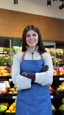 Kollarını kavuşturmuş gülümseyen bir kadın bakkalın portresi modern bir süpermarketin taze ürün reyonunda gururla duruyor. Mavi bir önlük giyiyor ve kameraya kendinden emin bir şekilde bakıyor.