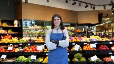 Kollarını kavuşturup kameraya gülümseyen, modern bir markette meyve ve sebze bölümünün önünde gururla duran genç ve güzel bir kadın işçinin portresi.