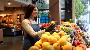 Gülümseyen, önlük ve eldiven takan genç bir manav modern bir süpermarkette veya yerel çiftçi pazarında taze, renkli organik meyveler sergiliyor.
