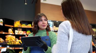 Önlük ve eldiven giyen arkadaş canlısı kadın bakkal bir kadına alışverişinde yardımcı olmak için dijital tablet kullanıyor ve üretim bölümünde mükemmel müşteri hizmetleri sunuyor.