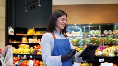 Önlük ve eldiven takan gülümseyen manav, bir marketin meyve ve sebze reyonunda bilgi sağlamak için dijital tablet kullanarak kadın müşteriye taze domates seçmesinde yardımcı olur.