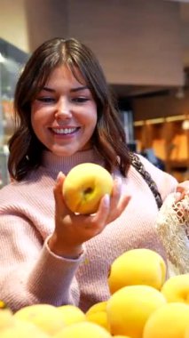 Yeniden kullanılabilir ağ torbasıyla neşeli genç bir kadın marketteki bir çöp kutusundan taze, olgun, sarı şeftalileri dikkatlice seçiyor, marketten alışveriş yaparken sağlıklı ve sürdürülebilir bir yaşam tarzını destekliyor.