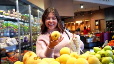 Gülümseyen kadın müşteri, süpermarketin üretim bölümünden olgun sarı elmaları seçiyor ve sağlıklı, sürdürülebilir bir yaşam tarzı için onları yeniden kullanılabilir bir ağ torbasına koyuyor.